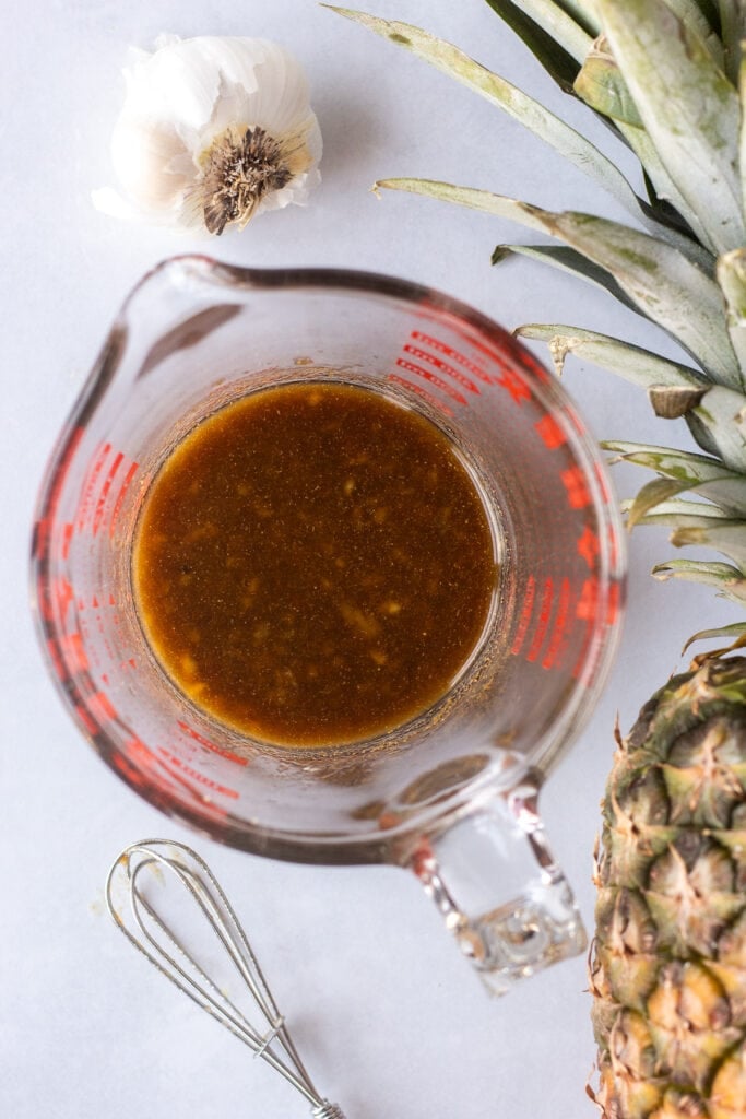 Top down shot of a measuring cup with a brown marinade in it. A garlic head, a pineapple, and a small whisk are off to different sides of the cup.