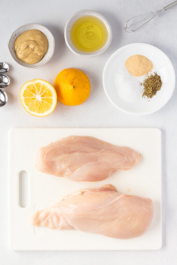 Top down shot of ingredients for grilled dijon chicken breast recipe, including raw boneless skinless chicken breasts, lemon, spices, dijon mustard, and oil.