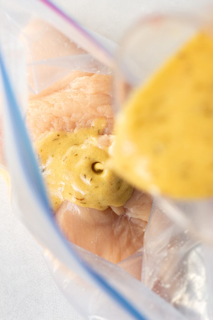 Pouring a dijon marinade into a ziploc bag with raw boneless skinless chicken breasts in it.