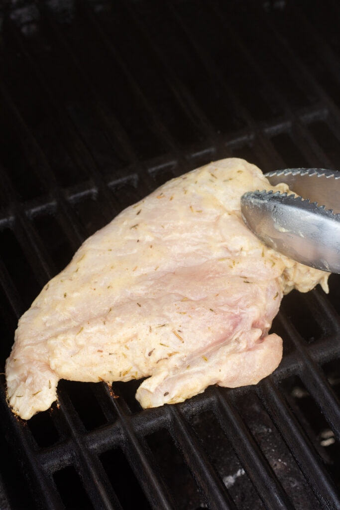 Raw marinated chicken breast being placed on a hot grill with metal tongs.