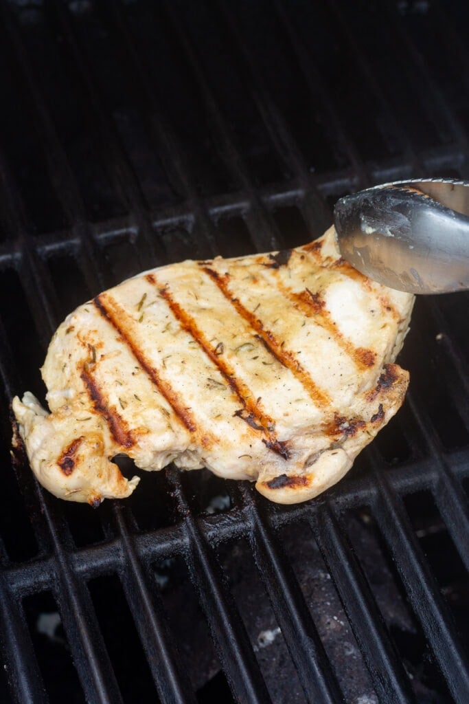 Tongs flipping over a grilled chicken breast on grill grates.