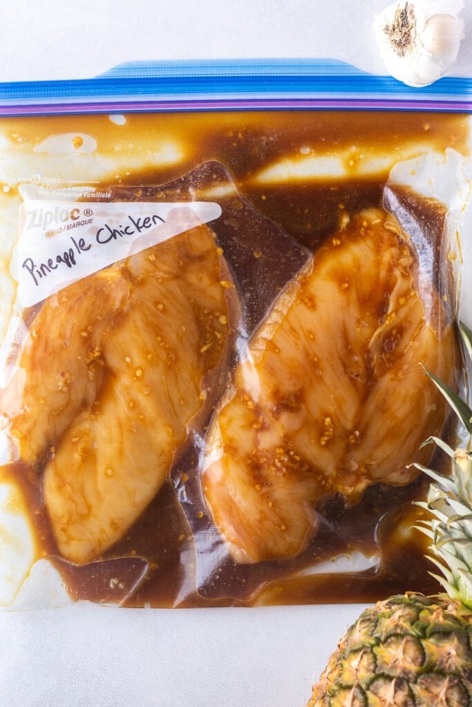 Top down shot of a sealed Ziploc bag with two raw boneless skinless chicken breasts marinating in a brown sauce. A head of garlic and a pineapple are to the side of the bag.