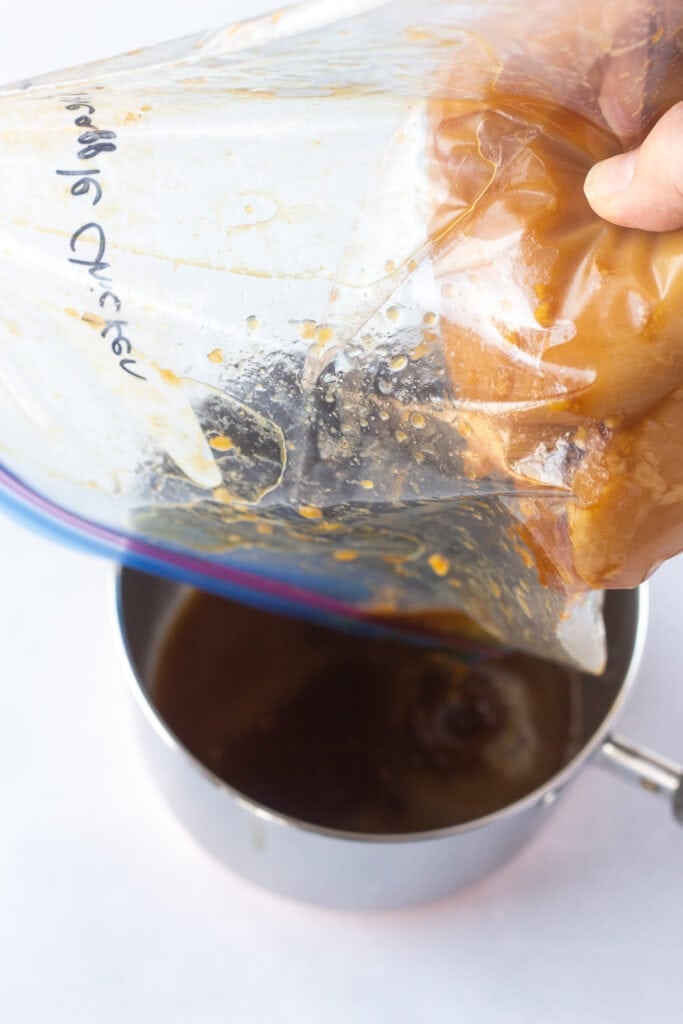 A hand holding a plastic ziploc bag with raw marinated chicken in it, pouring out the marinade into a small saucepan.