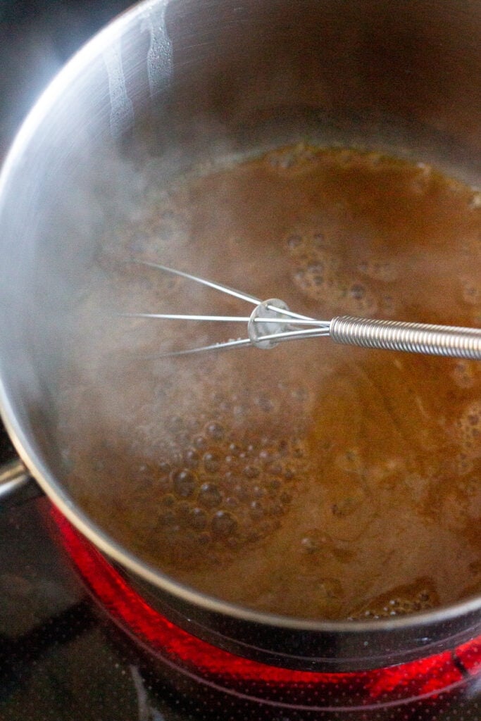 Close up of used pineapple marinade boiling in a small saucepan on the stove. The saucepan as a whisk in it and there is steam coming out of the pot.