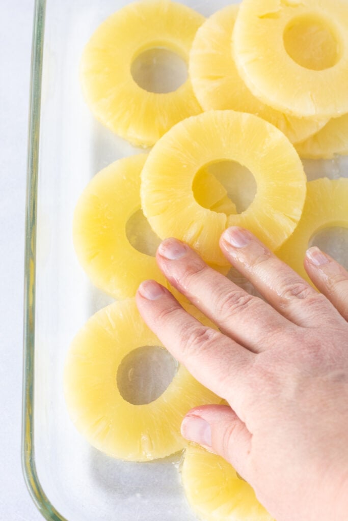 A hand rubbing oil on canned pineapple slices that are in a glass baking dish.