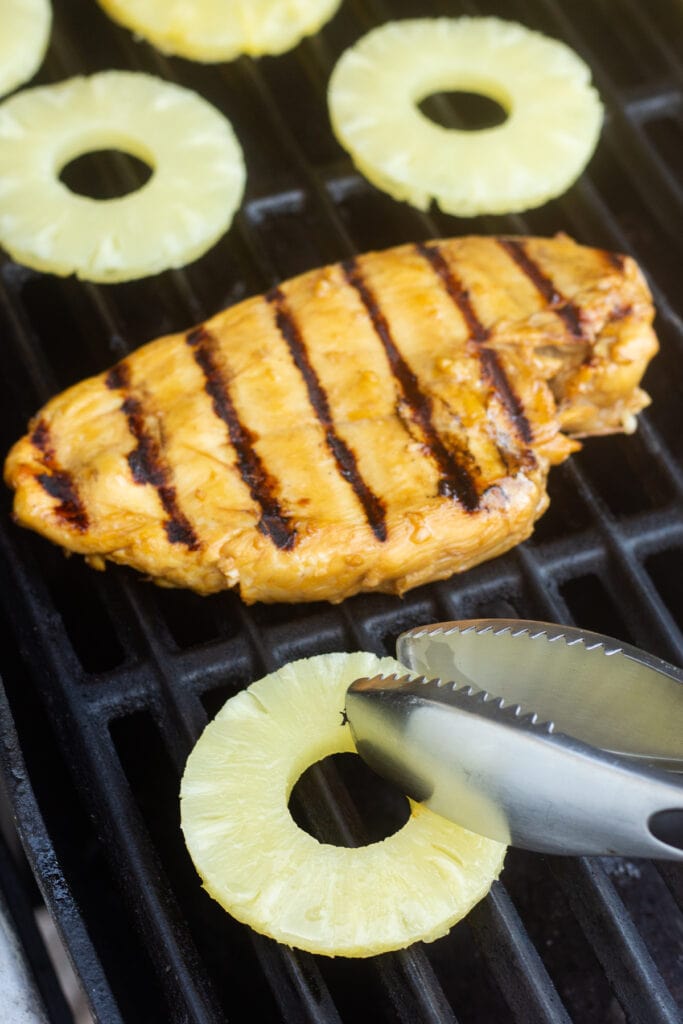 A pair of tongs placing canned pineapple rings onto a hot grill with a chicken breast grilling next to it.