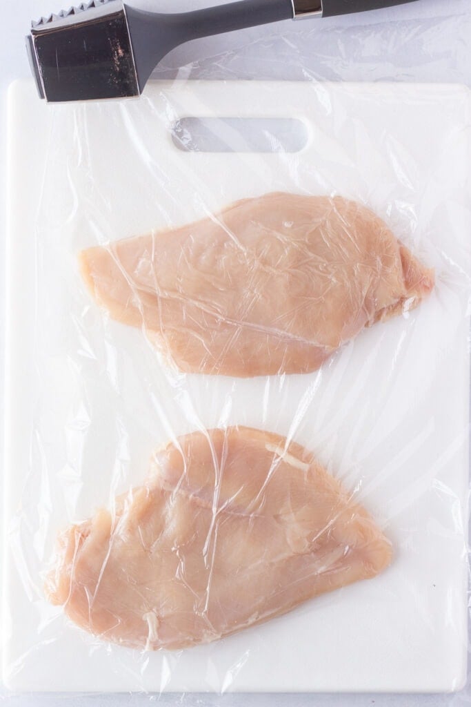 Top down shot of a pair of raw boneless skinless chicken breasts on a white cutting board covered by plastic wrap.