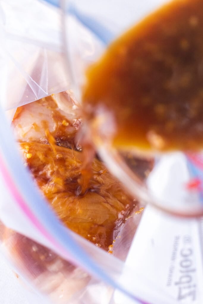 Close up of a brown marinade being poured out of a measuring cup into a ziploc bag with 2 raw boneless skinless chicken breasts in it.
