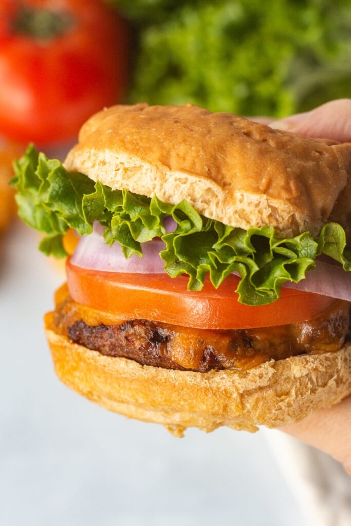 Close up of a hand holding a gluten free burger with a tomato and head of lettuce in the background.