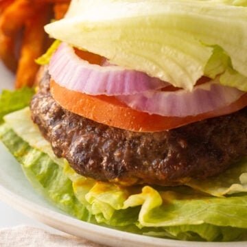 A close up of a paleo burger topped with tomato, red onion, and lettuce on a plate next to sweet potato fries.