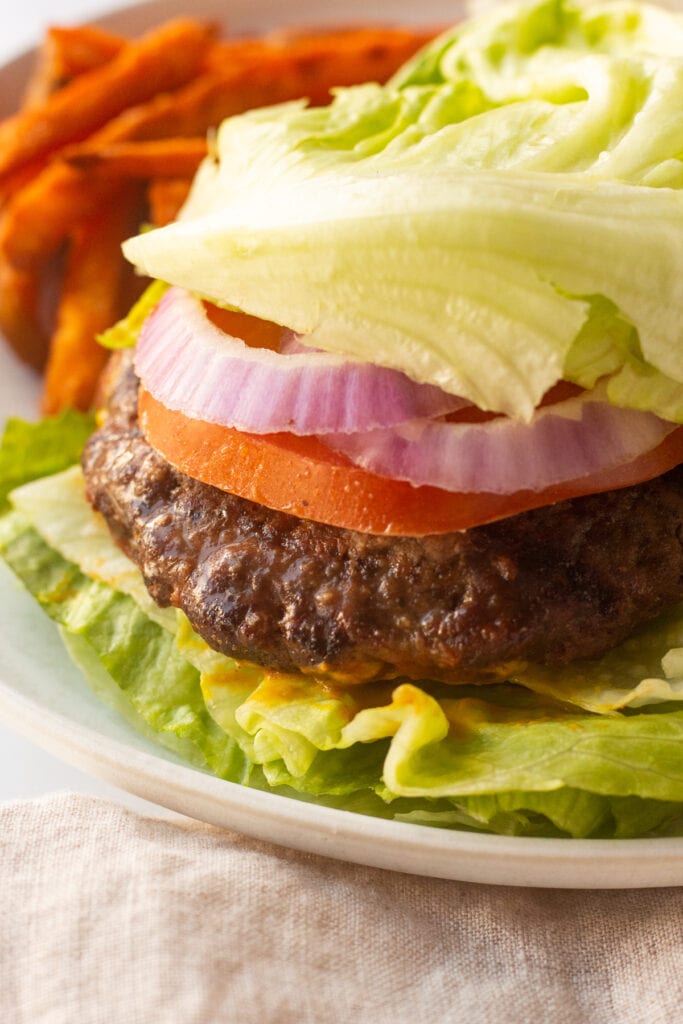 A close up of a paleo burger topped with tomato, red onion, and lettuce on a plate next to sweet potato fries.