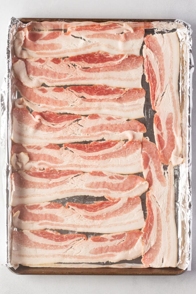 Top down shot of raw bacon in a single layer on a foil-lined rimmed baking sheet.
