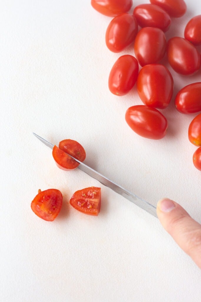 Close up of cherry tomatoes being quartered on a white cutting board.
