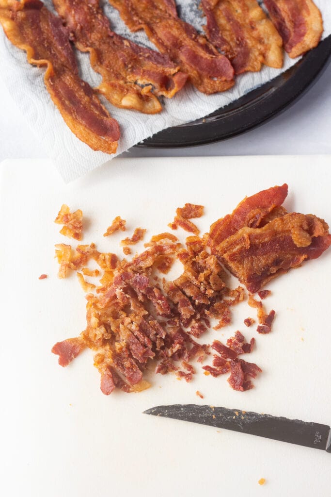 Strips of cooked bacon being cut into small pieces on a white cutting board.