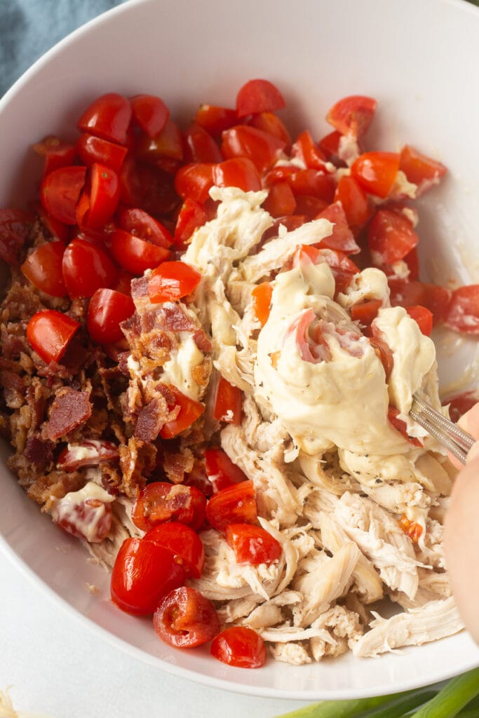 A spoon mixing mayo, bacon, chicken, pepper, and tomatoes together in a white bowl.