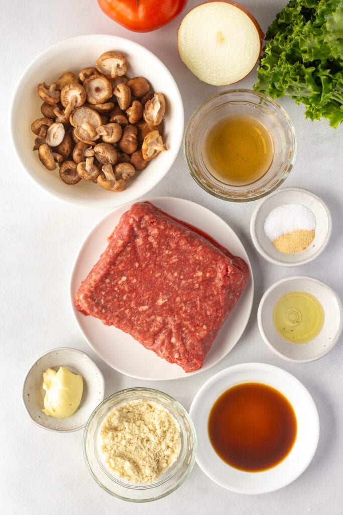 Top down shot of ingredients for a gluten free burger on a white background, including ground beef, spices, mushrooms, onion, and mayo.