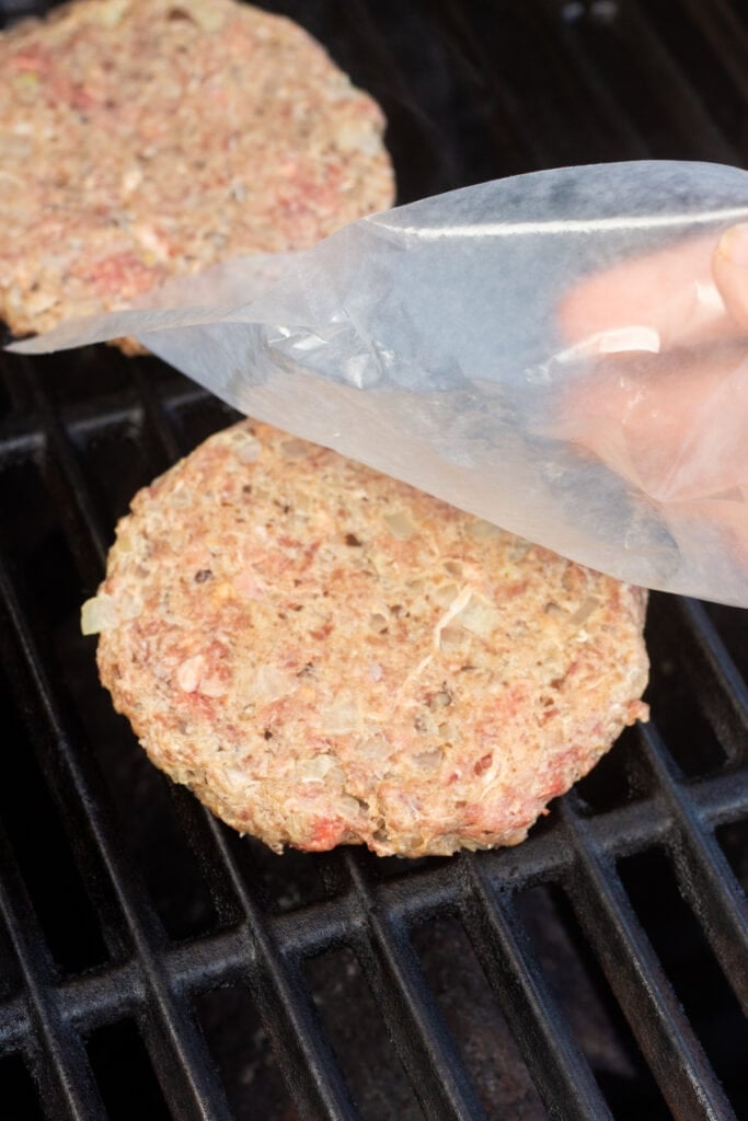A hand placing a raw burger patty on a grill and taking off the wax paper it was on.