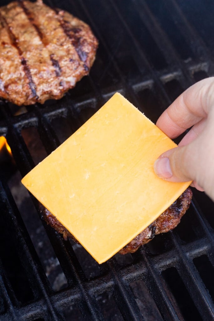 A hand placing a piece of sliced cheddar cheese on a cooked burger patty on a grill.