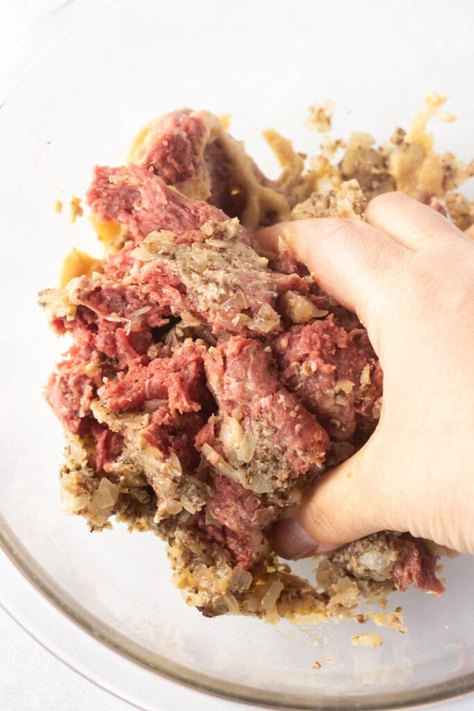 Close up of a hand mixing ground beef with sauteed mushrooms and onions and seasonings together in a clear bowl.