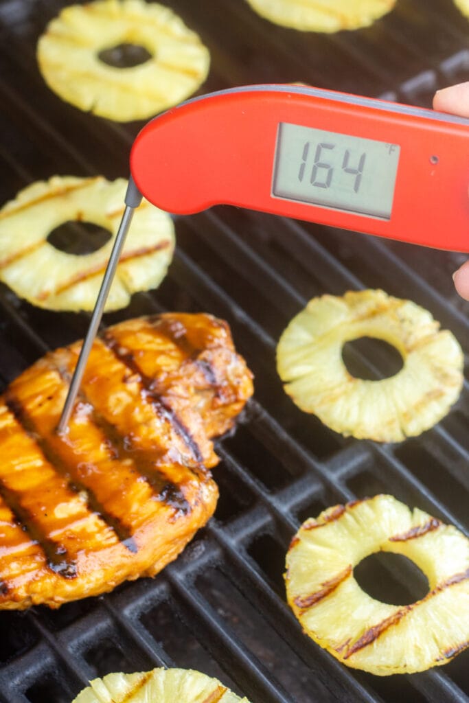 A red meat thermometer sticking into a grilled chicken breast that reads 165F. The chicken is surrounded by slices of grilled pineapple on the grill as well.