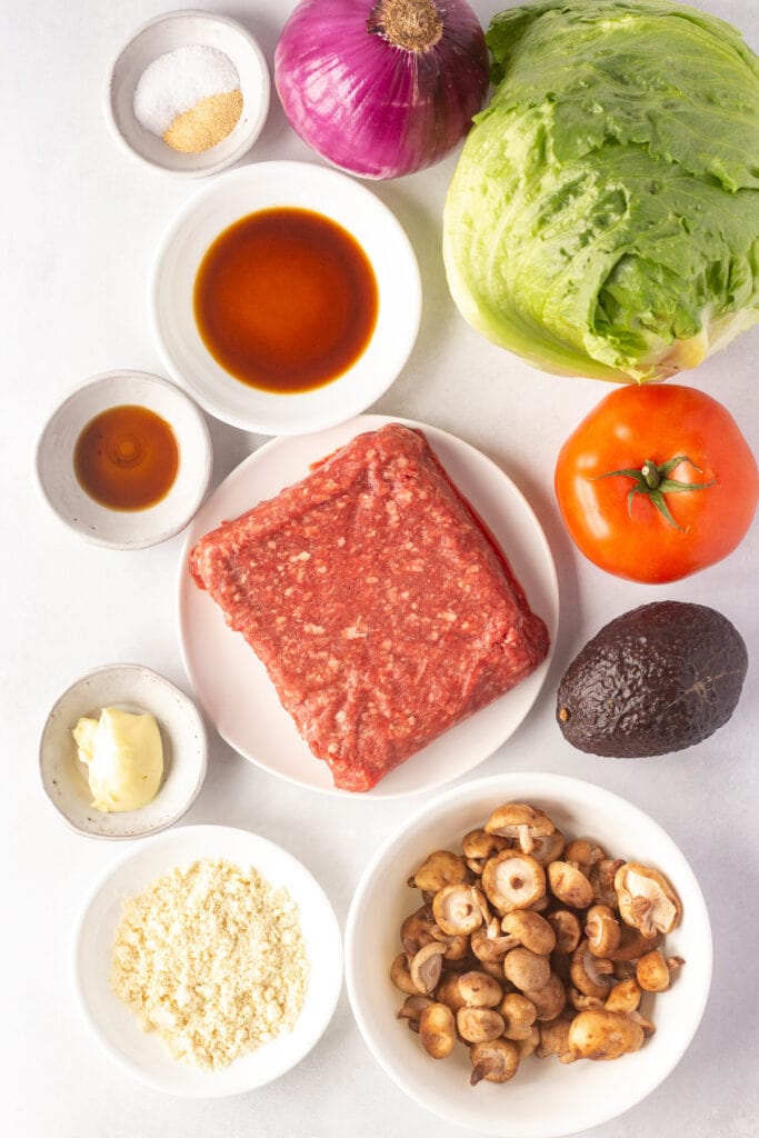 Top down shot of ingredients for paleo burgers, including ground beef, shiitake mushrooms, sauces and seasonings, red onion, lettuce, tomato, and avocado.