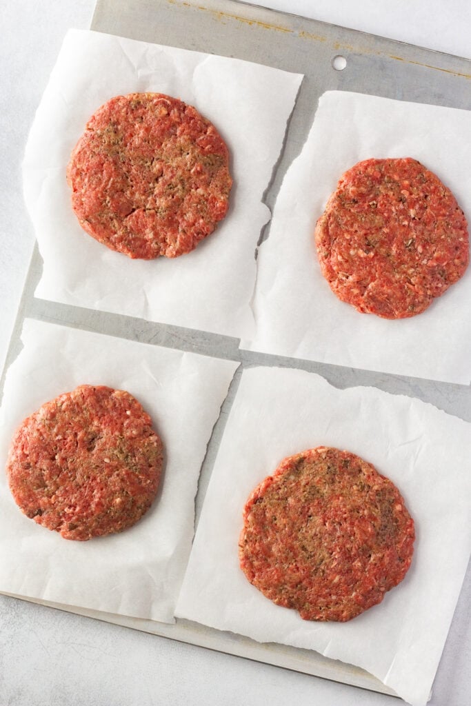 Top down shot of 4 uncooked hamburger patties on individual pieces of white wax paper on top of a cooking sheet.