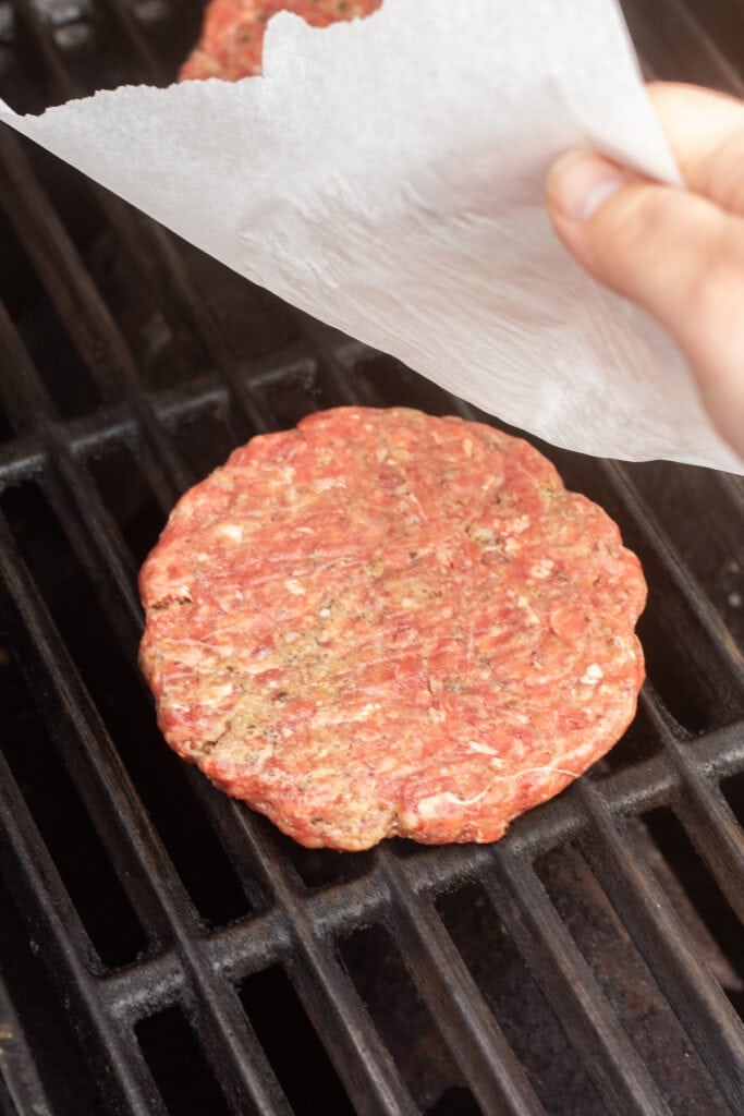 A hand removing the wax paper off an uncooked hamburger patty that's been placed on a hot grill.