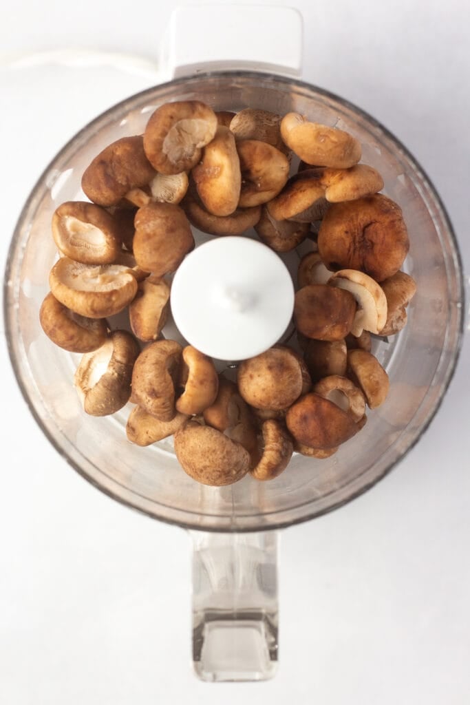 top down shot of shiitake mushroom caps in a small food processor on a white background.