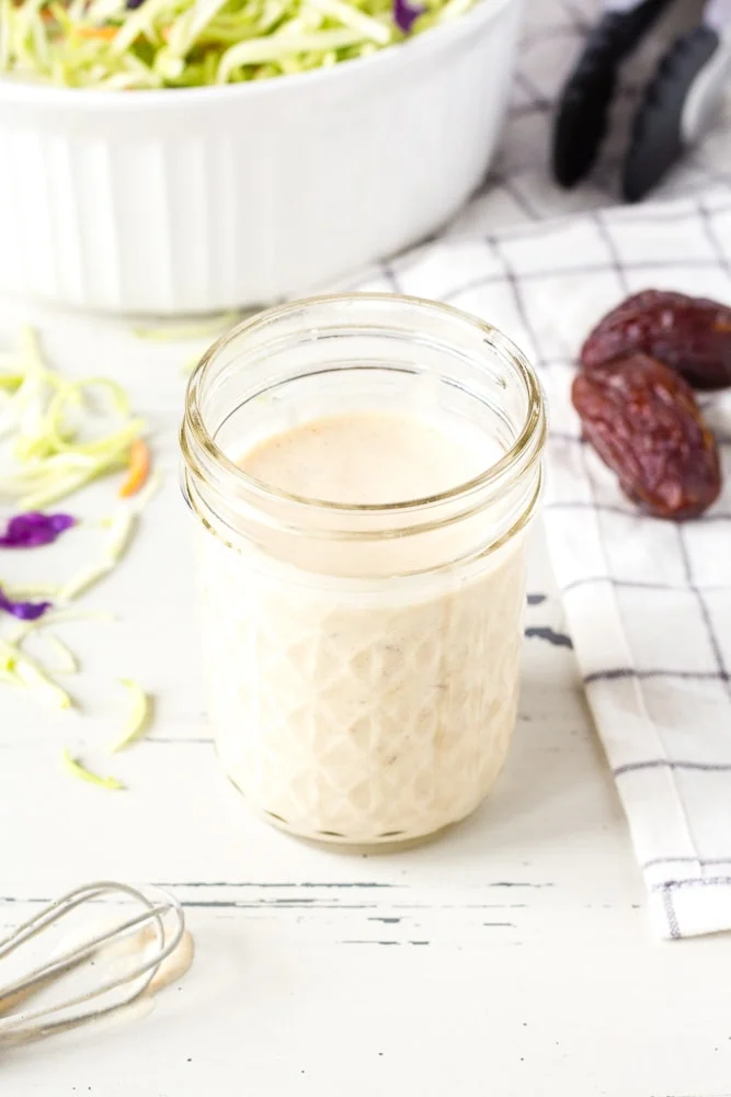 creamy homemade coleslaw dressing in a small mason jar next to a bowl with slaw in it.