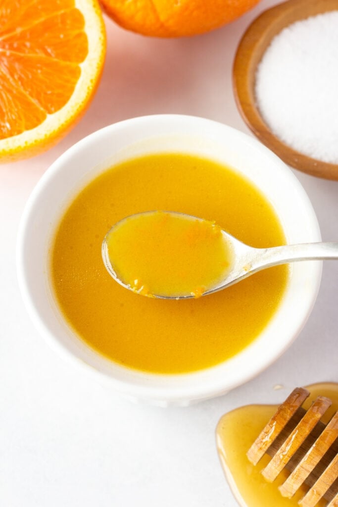 A white bowl with orange dressing in it, with a spoon dipping a bit of dressing out. It's surrounded by a half an orange, a small wood bowl with kosher salt, and a honey stick dripping with honey.
