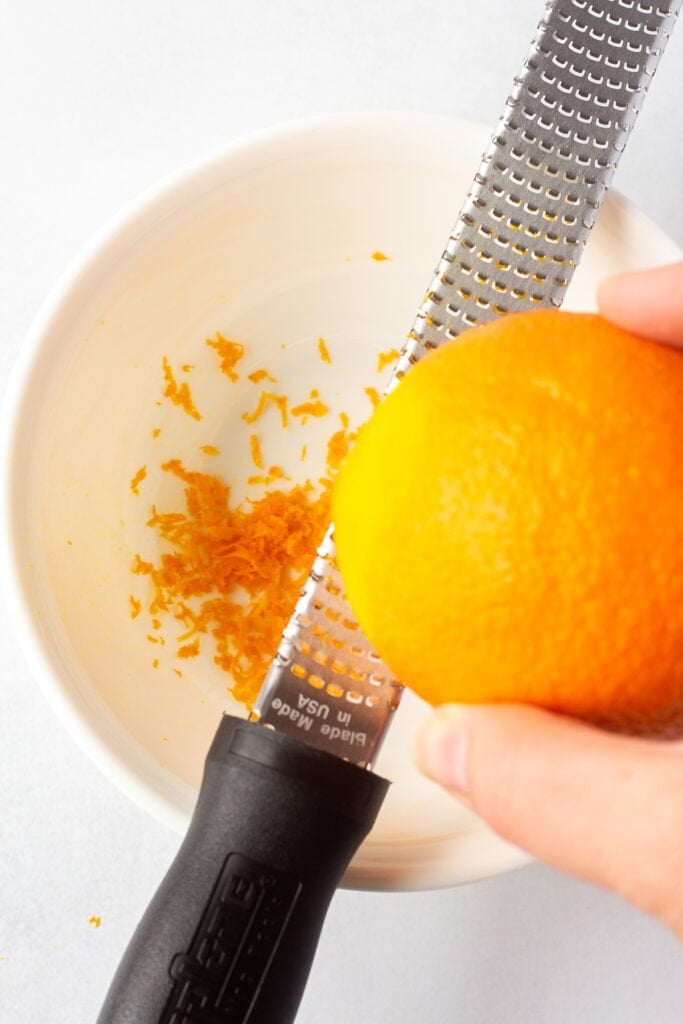 Close up of zesting an orange with a microplane over a medium white bowl.