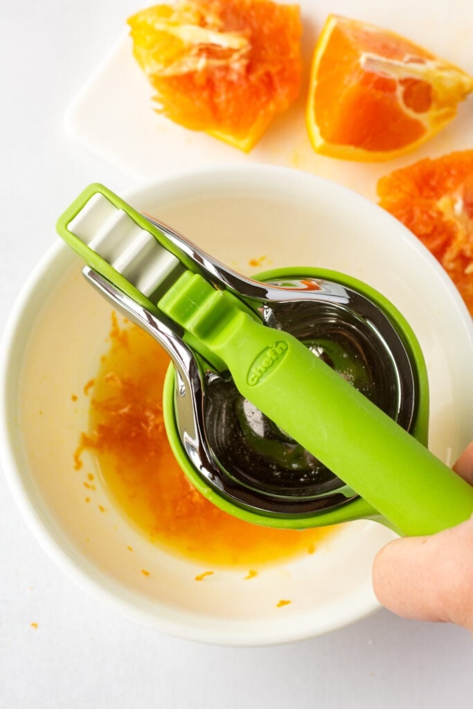 Juicing pieces of orange with a green handheld juicer over a medium white bowl.