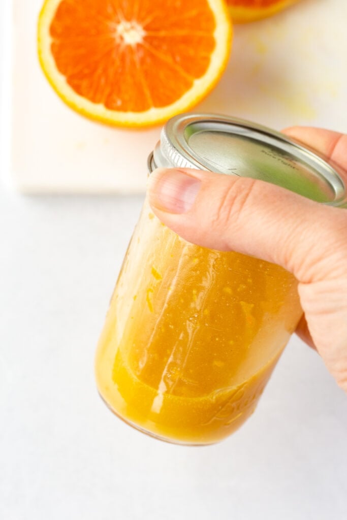 A hand holds a small mason jar with a lid containing orange dressing. Half an orange on a white cutting board is in the background.