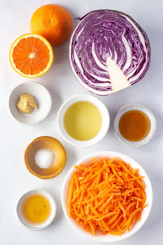 Top down shot of ingredients for purple cabbage and carrot coleslaw in white bowls, including half a purple cabbage and a cut orange, all on a white background.