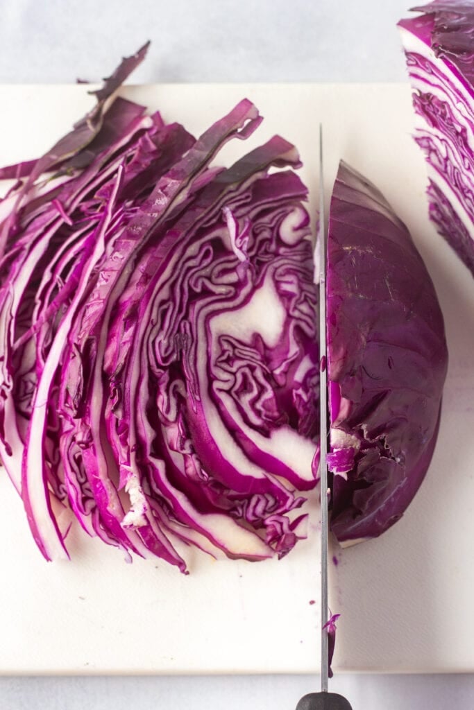 A quarter of a head of purple cabbage being cut into thin slices with a knife on a white cutting board.
