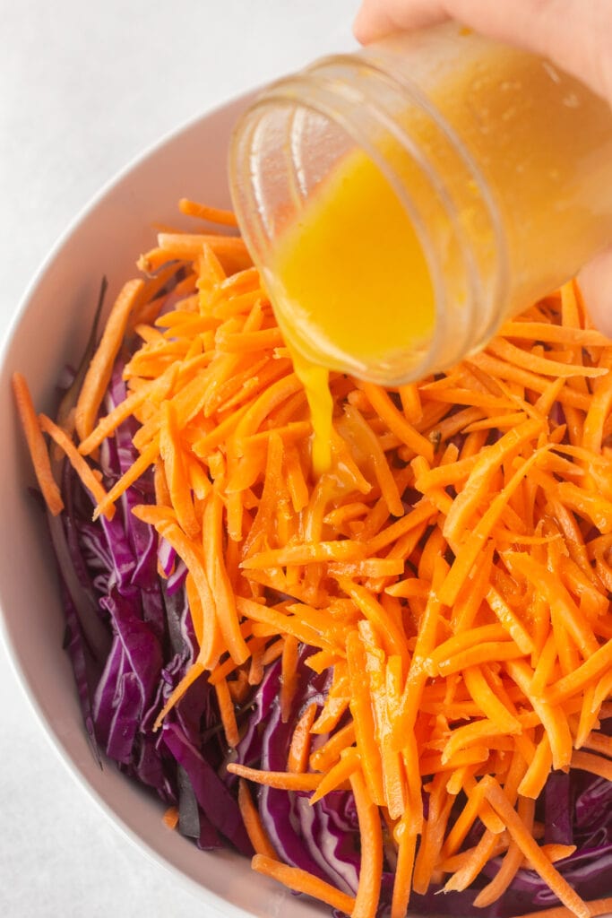 Pouring orange vinaigrette dressing into a white bowl filled with thinly sliced purple cabbage and carrot matchsticks.