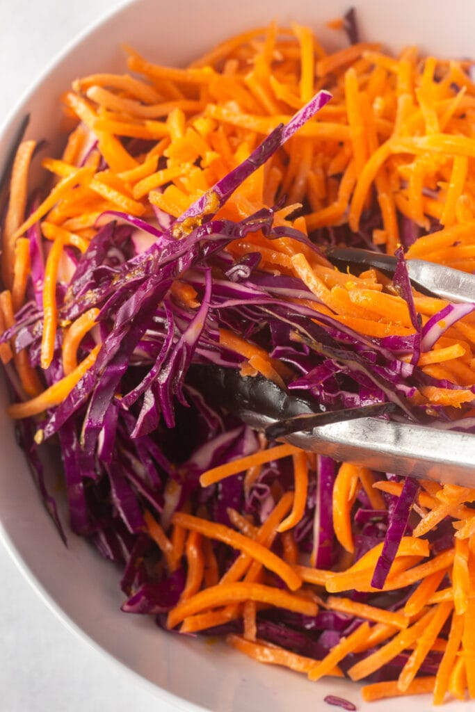 Close up of a pair of tongs mixing up shredded carrots and sliced purple cabbage with a vinaigrette dressing in a white bowl.