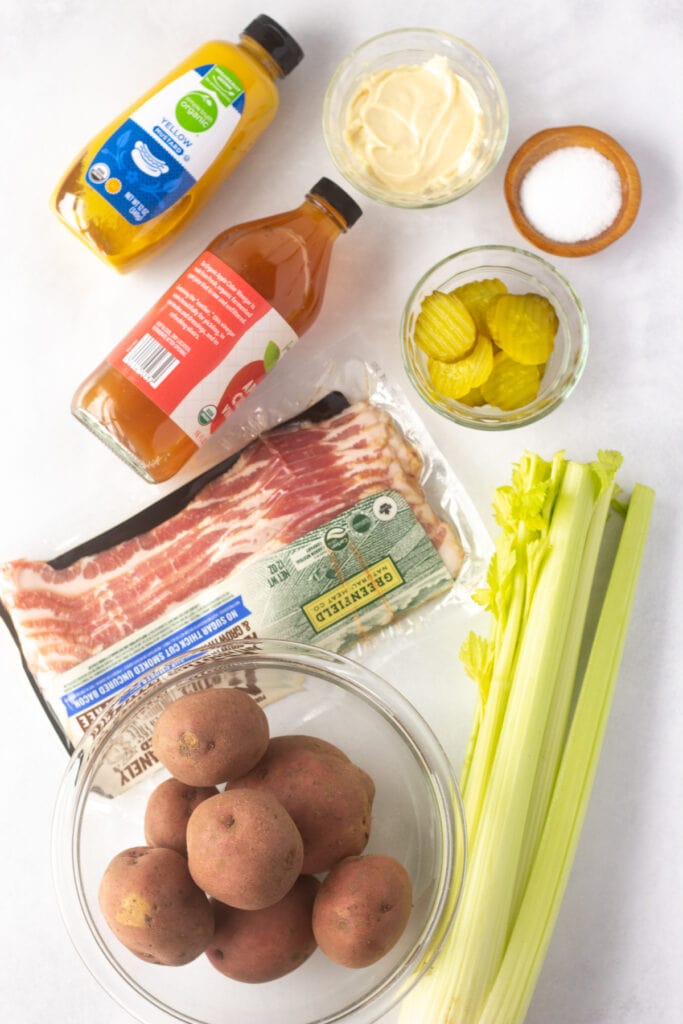 Top down shot of ingredients for red skin potato salad on a white background, including red potatoes, celery, pickles, mustard, mayo, bacon, and apple cider vinegar.