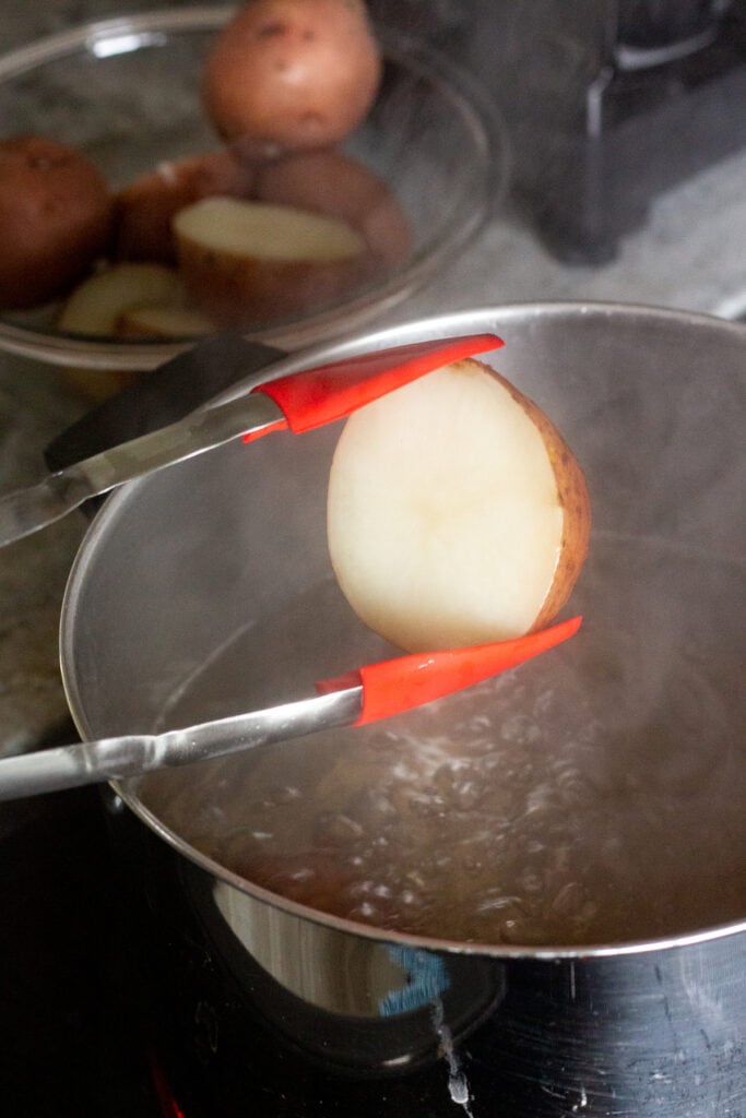 Red tongs removing cooked potato from a pot of boiling water on a stovetop.