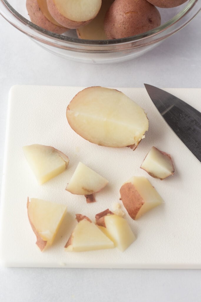 Cooked red potato being cut into smaller pieces on a white cutting board with a large knife.