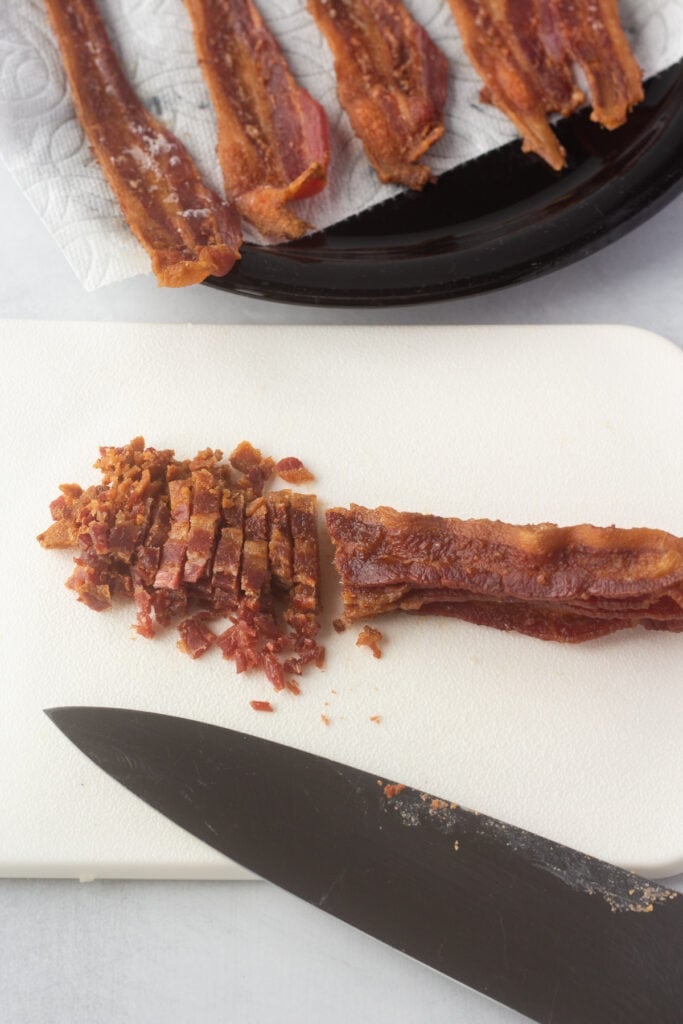 Close up of cooked bacon strips being cut into small pieces on a white cutting board with a large knife.