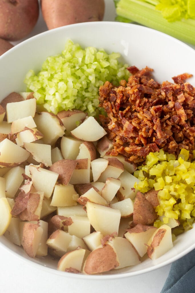 A large white bowl with cut potatoes, celery, bacon, and dill pickle in it.