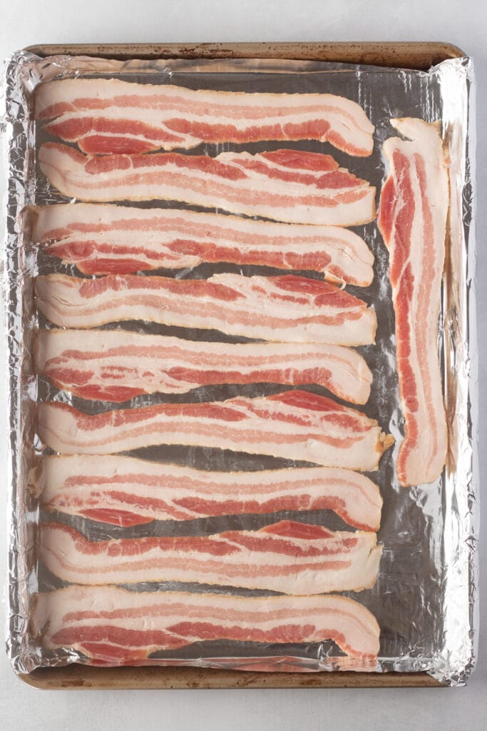 Top down shot of a rimmed back sheet lined with foil with strips of raw bacon on it in a single layer.