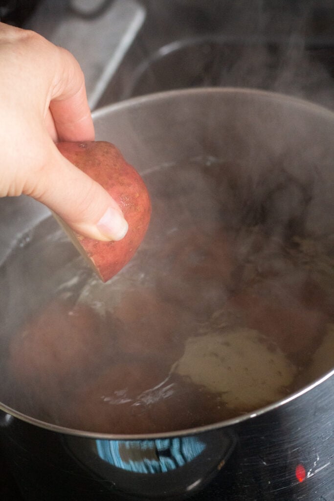 Close up of a hand placing raw cut red potatoes into a pot of boiling water on a stovetop.