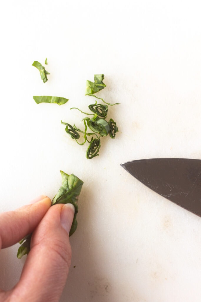 Close up of fresh basil leaves rolled together and being cut into small ribbons on a white cutting board.