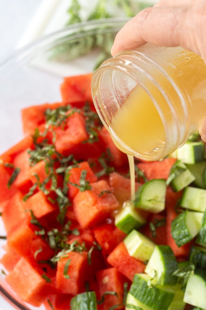 Close up of golden salad dressing being poured over cut watermelon, chopped cucumber, and ribbons of fresh basil in a large bowl.