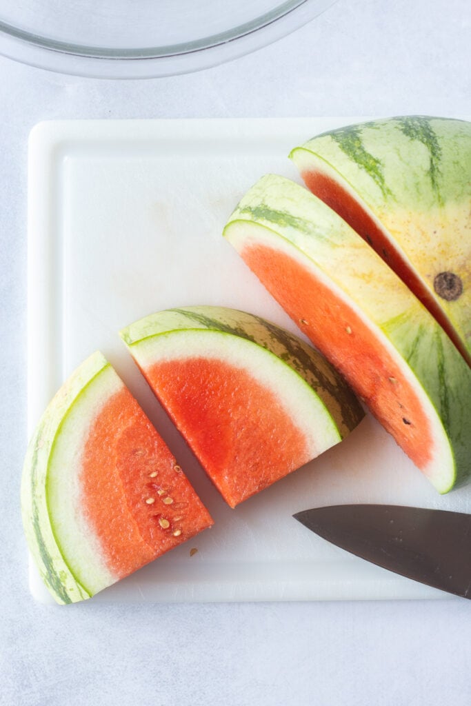 Top down shot of half a watermelon being sliced into quarters on a white cutting board.
