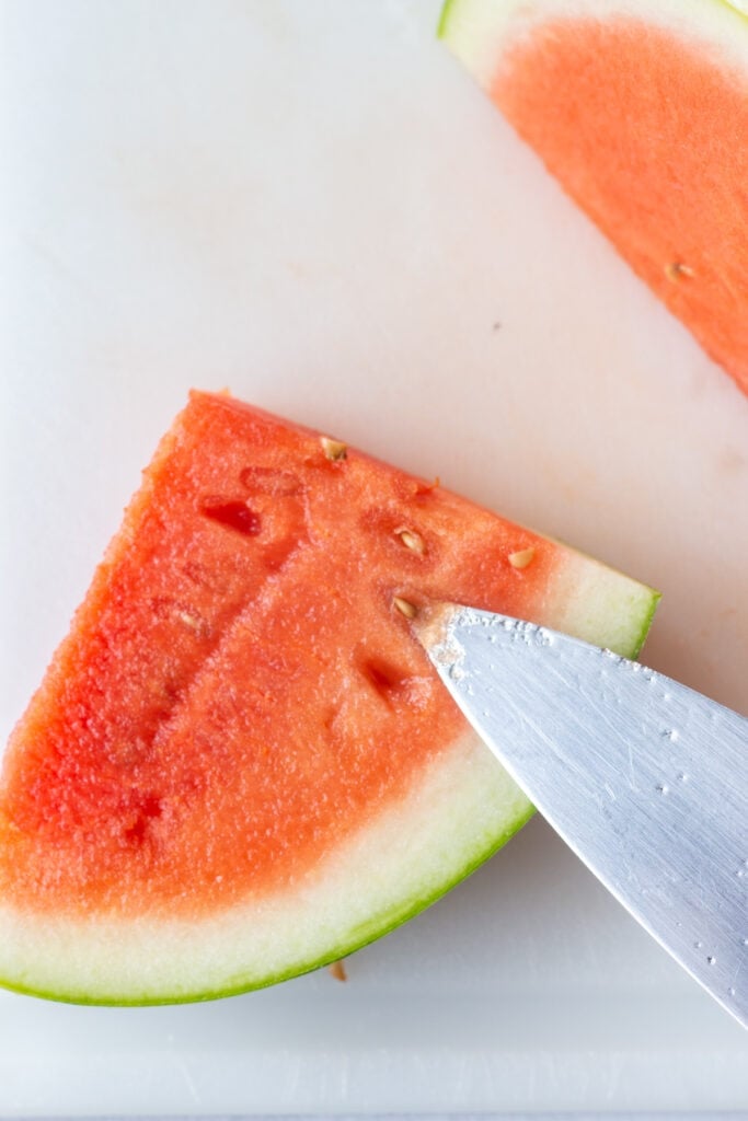 Close up of a sharp knife digging seeds out of a quartered slice of a watermelon.