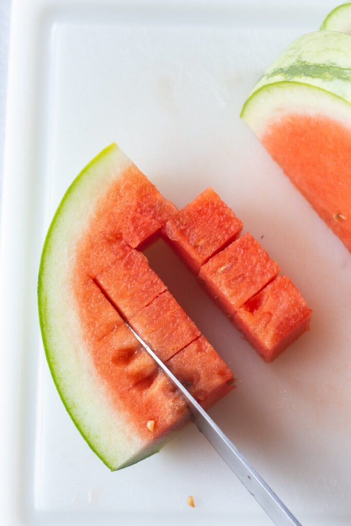Close up of a quartered slice of a watermelon being cut into cubes by a large knife, all on a white cutting board.