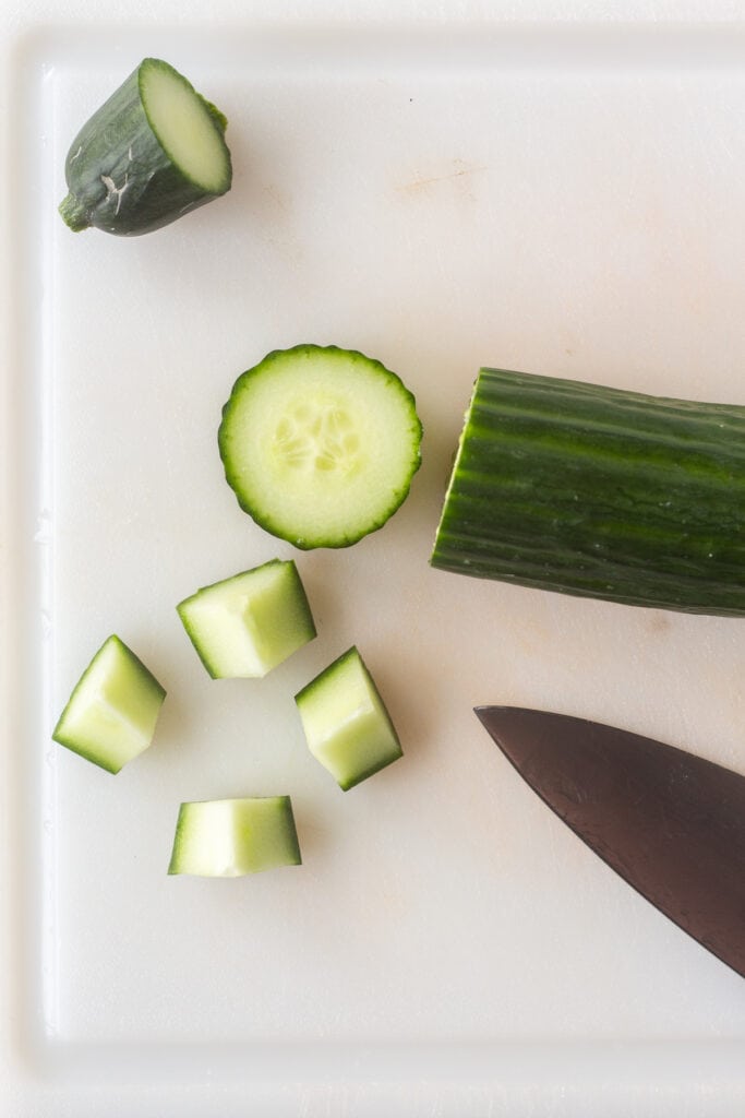 A cucumber being sliced and chopped into quarters on a white cutting board.